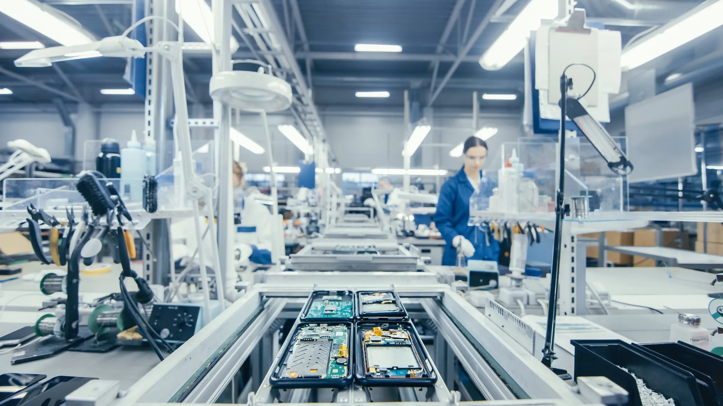 Eine Montagelinie in einer Elektronikfabrik, Geräteplatinen in einem Träger auf dem Förderband, dahinter eine Arbeiterin an der Linie.